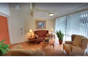 A cozy living room area with a red loveseat, two beige armchairs, a wooden coffee table with a floral centerpiece, two side tables with lamps, a framed picture on the wall, and large vertical blinds covering the windows letting in natural light.