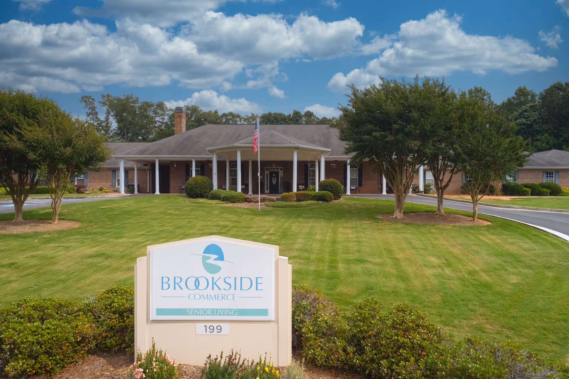 Front exterior of Brookside Commerce senior living facility with entrance, flagpole, and a sign on a manicured lawn.