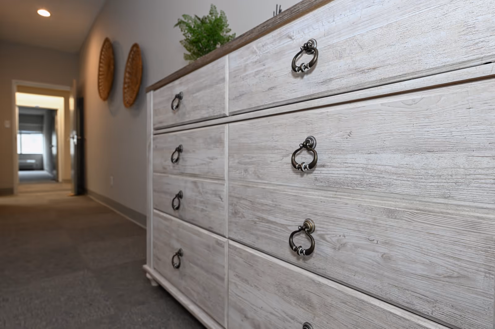 A light wood dresser with metal ring handles sits in a carpeted hallway leading to an open doorway.