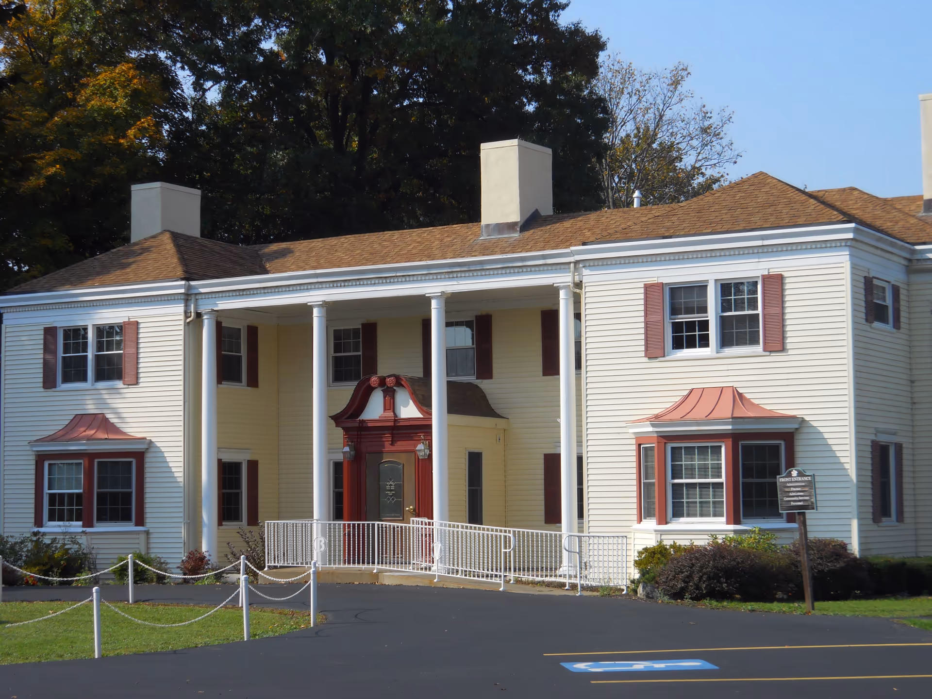 Front exterior view of a two-story senior living facility building with cream-colored siding, red window shutters, and a red front door with a decorative awning. The entrance is supported by four white columns and has a wheelchair ramp. There is a parking lot with a handicap parking space in front and trees in the background.
