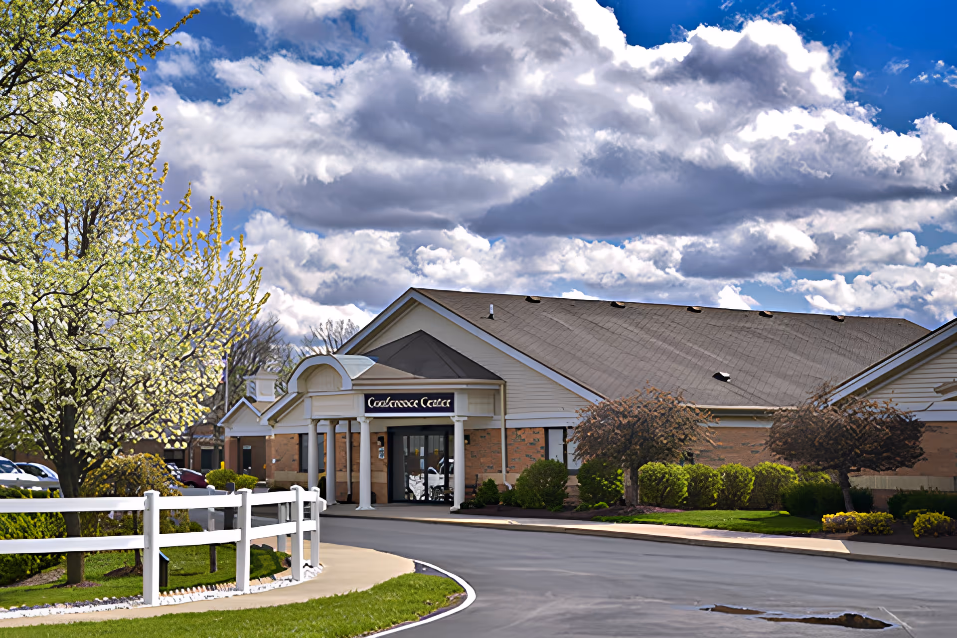 Exterior view of a single-story brick building with a peaked roof and white columns at the entrance. The building is surrounded by well-maintained landscaping including green bushes, small trees, and a white fence. The sky is partly cloudy with patches of blue visible.