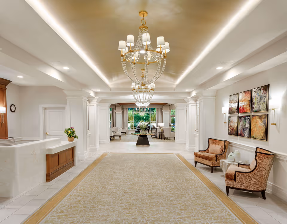 A spacious and elegant interior hallway of a senior living facility with a long patterned carpet, two large chandeliers hanging from the ceiling, a reception desk on the left, and two upholstered chairs with a small table on the right. The hallway leads to a sitting area with armchairs and a large window showing greenery outside. The walls are decorated with colorful abstract paintings and wall sconces.