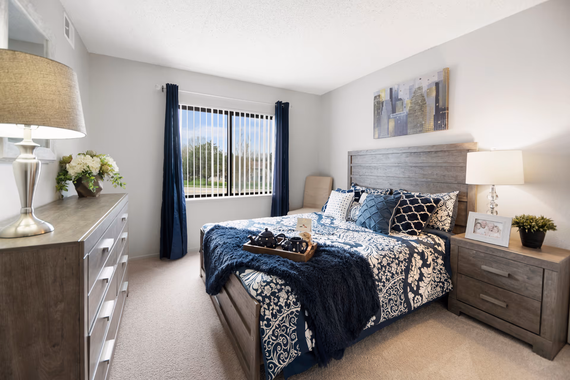 Bright furnished bedroom with a bed, wooden headboard, nightstand and dresser, a window with vertical blinds, and decorative pillows and lamps.