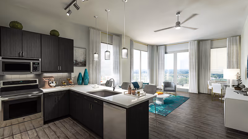 Modern open-concept living space featuring a kitchen with dark wood cabinets, stainless steel appliances, and a white countertop island with a sink. The living area includes a teal rug, a coffee table, armchairs, and large windows with white curtains allowing natural light to fill the room. A ceiling fan is mounted above, and a dining area with a white table and chairs is visible near the windows.