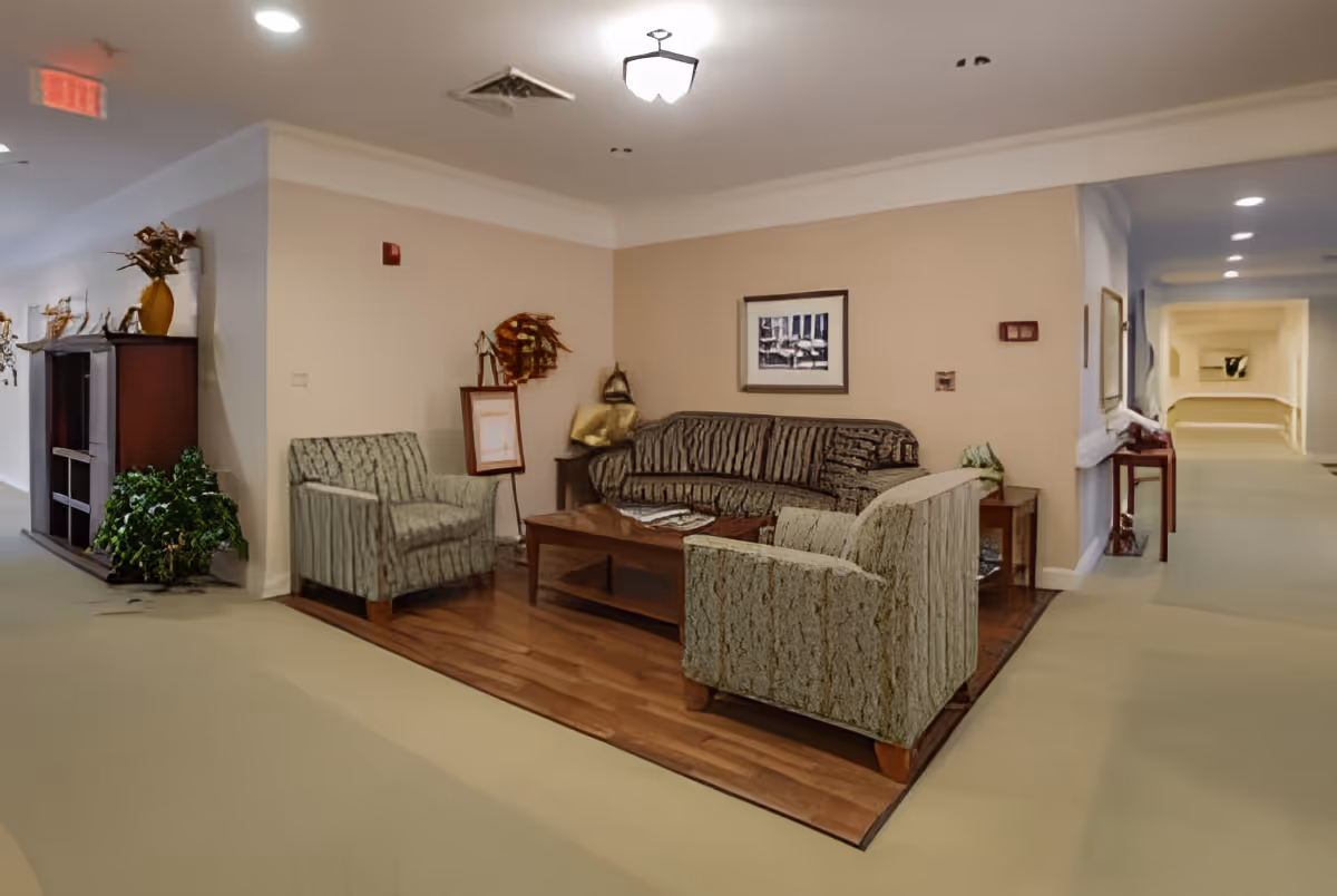 A cozy seating area in a senior living facility hallway featuring a patterned sofa, two matching armchairs, a wooden coffee table, and decorative items including framed artwork and plants. The seating area is set on a wooden floor section surrounded by carpeted flooring, with soft lighting from ceiling fixtures.