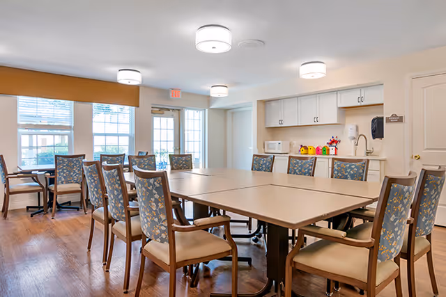 A bright and clean dining room with multiple tables pushed together to form a large square table surrounded by chairs with floral patterned upholstery. The room has large windows letting in natural light, a door leading outside, and a kitchenette area with cabinets, a microwave, and colorful decorative items on the counter.