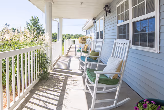 Covered front porch with white rocking chairs, cushions, and potted plants along a light blue building.