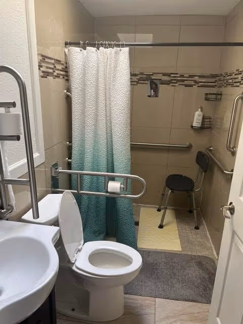 Accessible bathroom with a white toilet, a sink, and a shower area with a white and teal gradient shower curtain. The bathroom has beige tiled walls with a decorative horizontal tile strip. There are metal grab bars near the toilet and in the shower area, a black shower chair, and a bottle of soap or shampoo on a wall-mounted shelf.