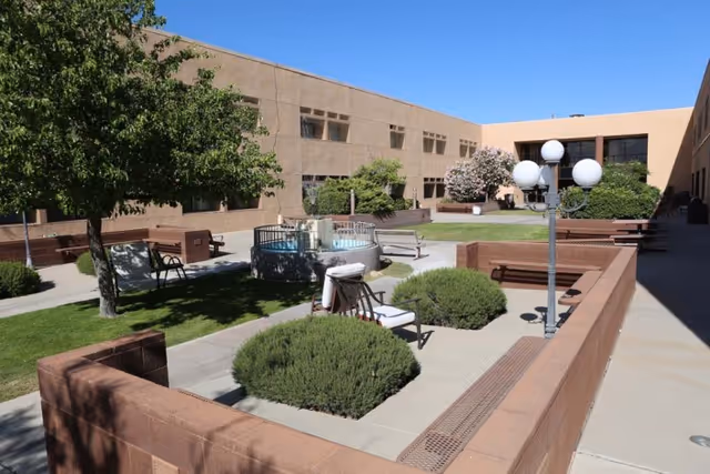 Outdoor courtyard area of a senior living facility with benches, a small circular fountain, green bushes, trees, and a lamp post with four globe lights, surrounded by a two-story beige building under a clear blue sky.