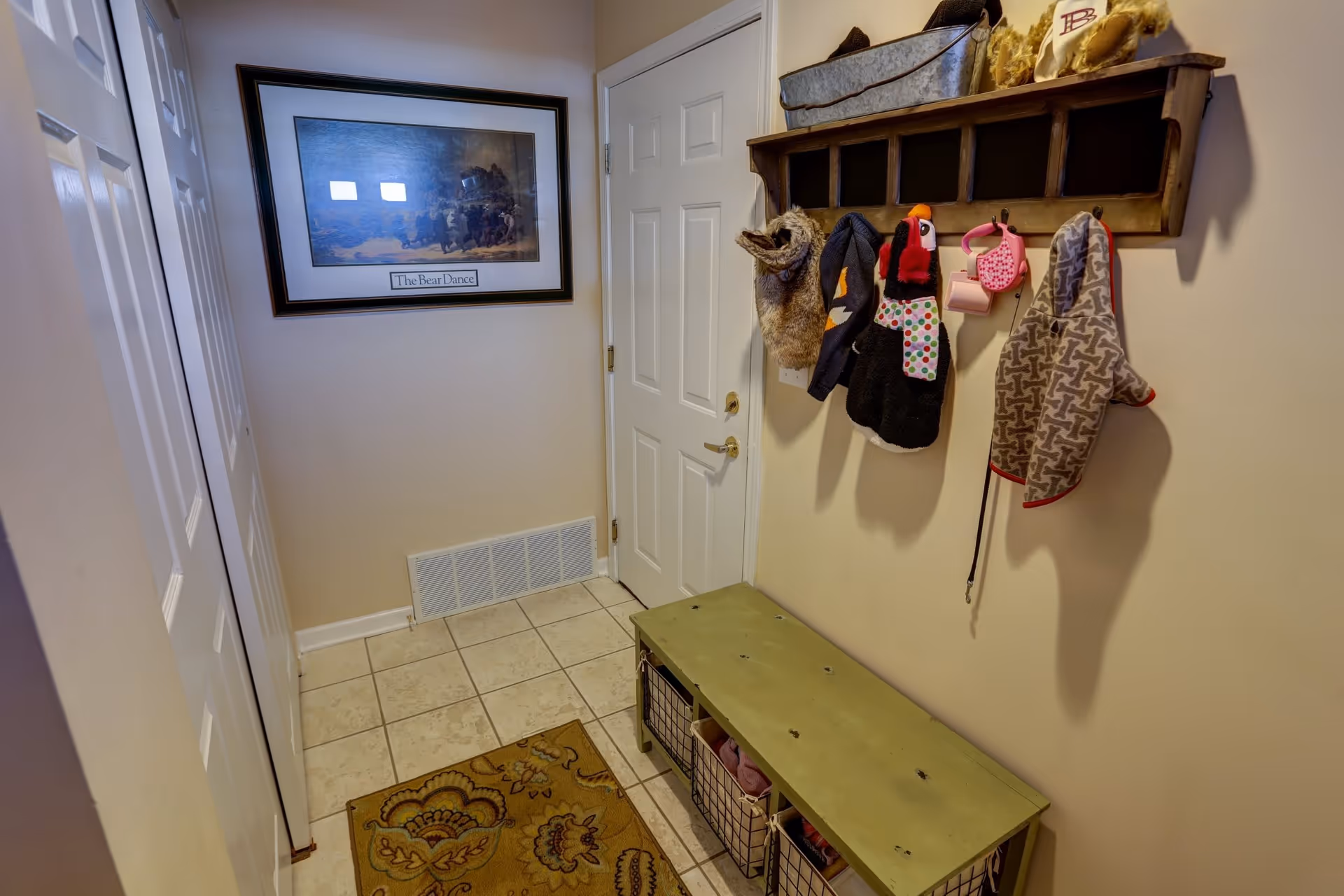 A small entryway with a white door, beige walls, and tiled floor. There is a green bench with wire baskets underneath on the right side, and above it, a wooden wall-mounted coat rack holding several dog jackets and accessories. A framed picture titled 'The Bear Dance' hangs on the wall opposite the door. A patterned rug is placed on the floor near the bench.