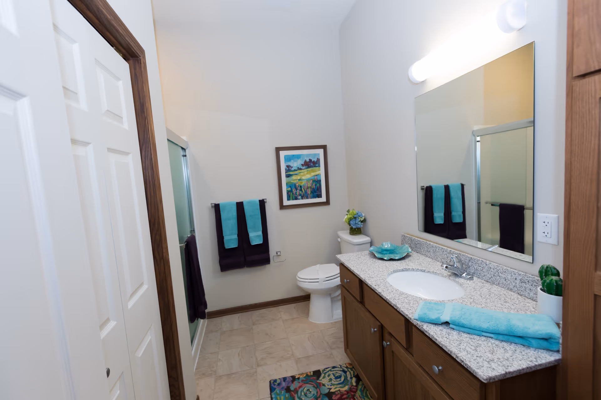 A clean bathroom with a granite-topped vanity and sink, large mirror, toilet, shower, and turquoise towels.