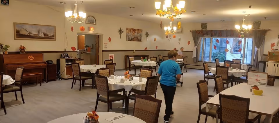 A dining room in a senior living facility decorated with autumn-themed decorations including leaves and pumpkins. Several round and rectangular tables are set with white tablecloths, cups, and small centerpieces. Two people are present, one walking away from the camera and another seated near the window. The room features chandeliers, a piano, and large windows with curtains.