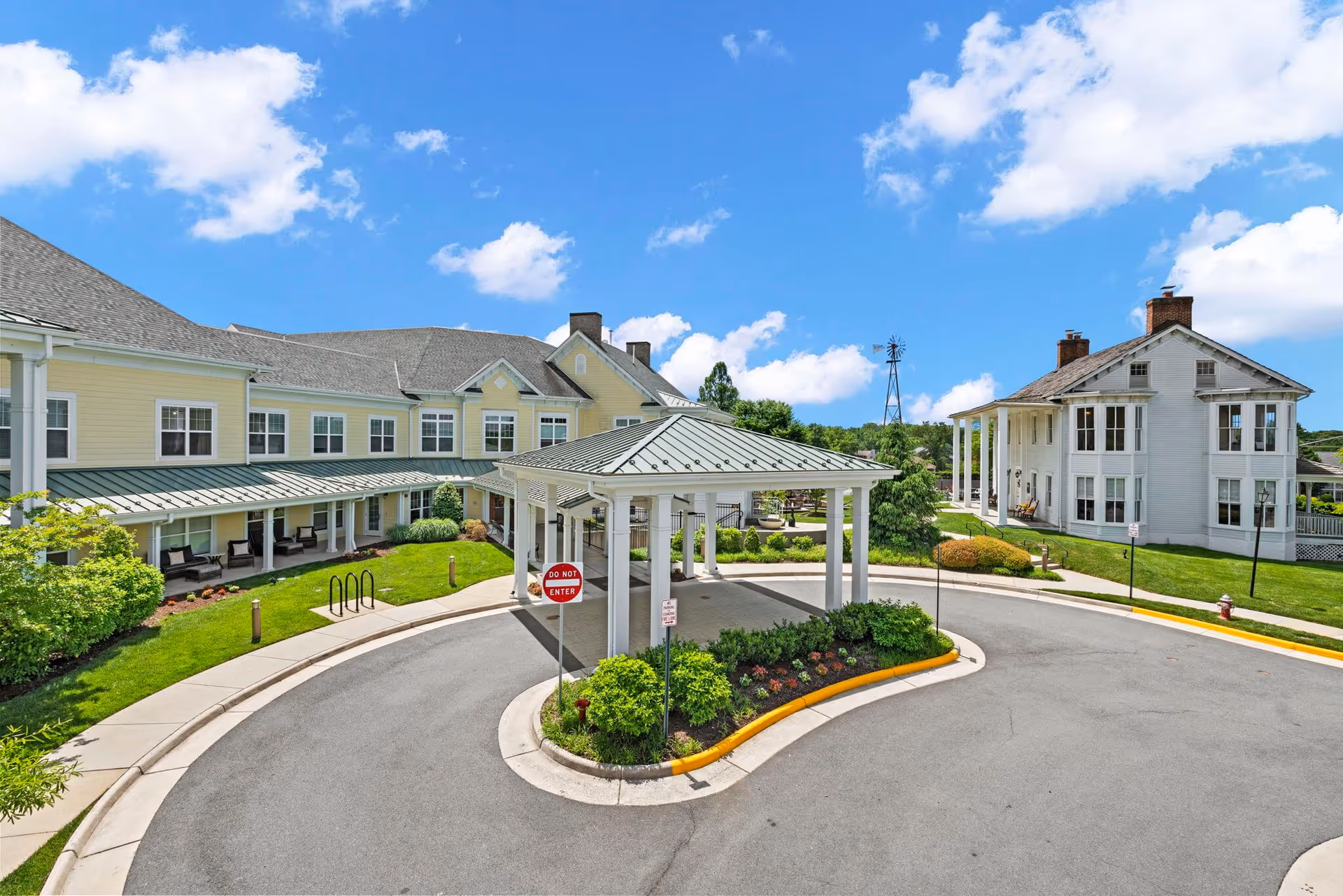 Front entrance and porte-cochere of a senior living facility with a landscaped circular driveway and two two-story buildings under a blue sky.