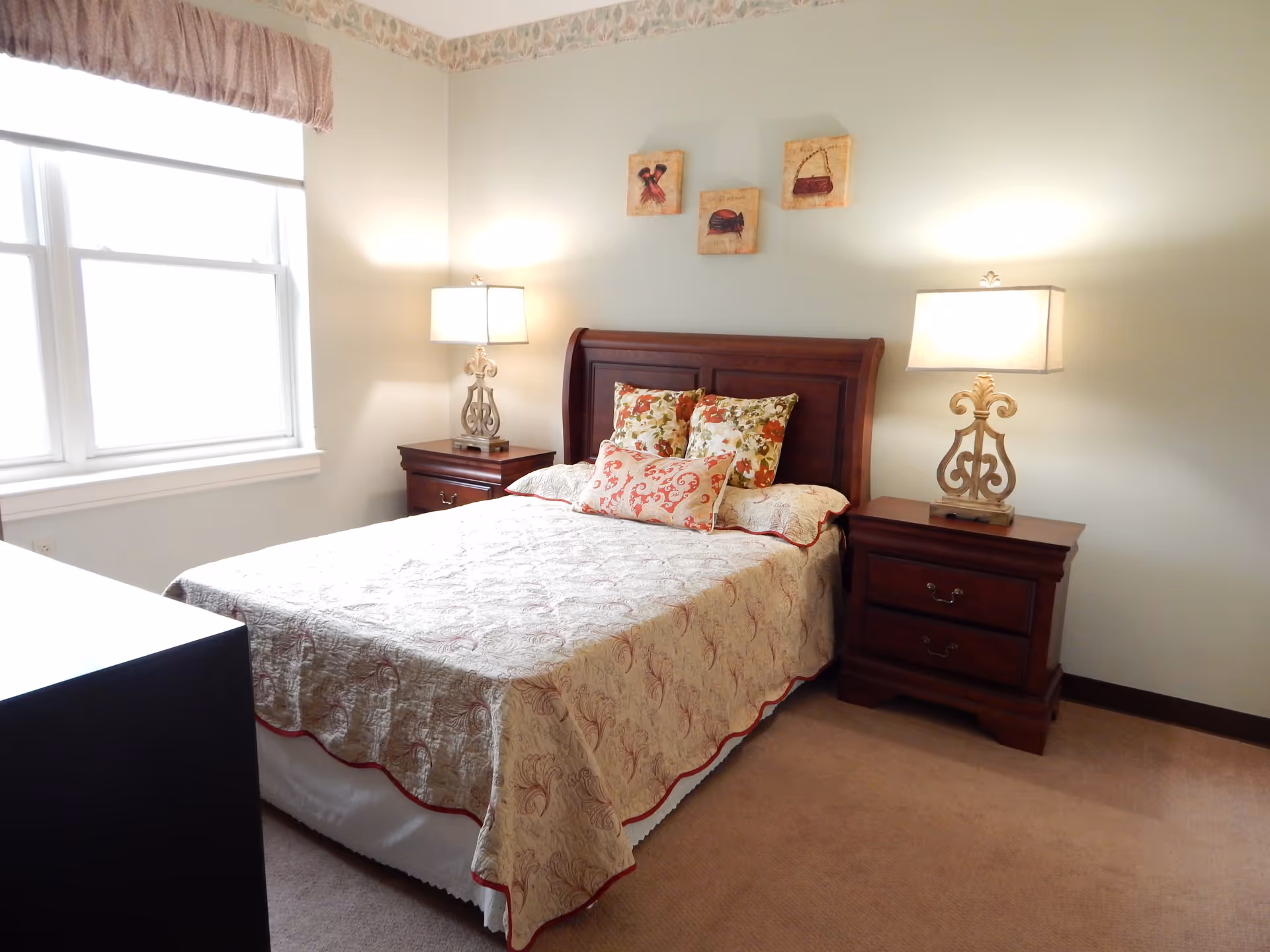 A bedroom with a double bed featuring a patterned bedspread and decorative pillows. There are two wooden nightstands on either side of the bed, each with a lamp. A window with a valance is on the left wall, and three small framed pictures hang above the headboard. The room has beige carpet and light-colored walls with a decorative border near the ceiling.