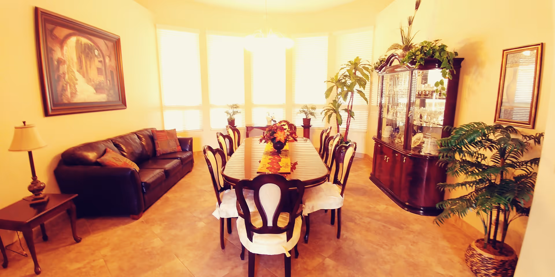A well-lit dining room with a long wooden dining table surrounded by eight chairs with white cushions. The table is decorated with a floral centerpiece and a colorful table runner. To the left, there is a dark brown leather sofa with cushions and a side table with a lamp. On the right side, there is a wooden china cabinet displaying glassware and decorative items, along with several potted plants. The room has large windows with blinds allowing natural light to fill the space, and a framed painting and mirror on the walls.