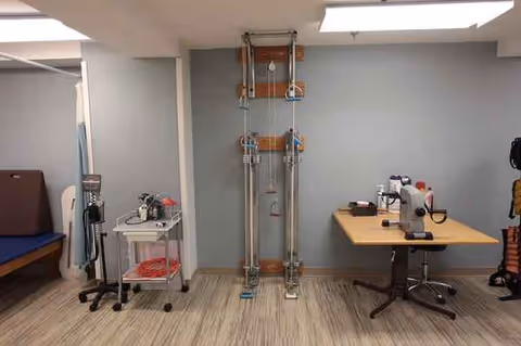 Clinical rehabilitation room with a standing frame, equipment cart, bed, and desk.
