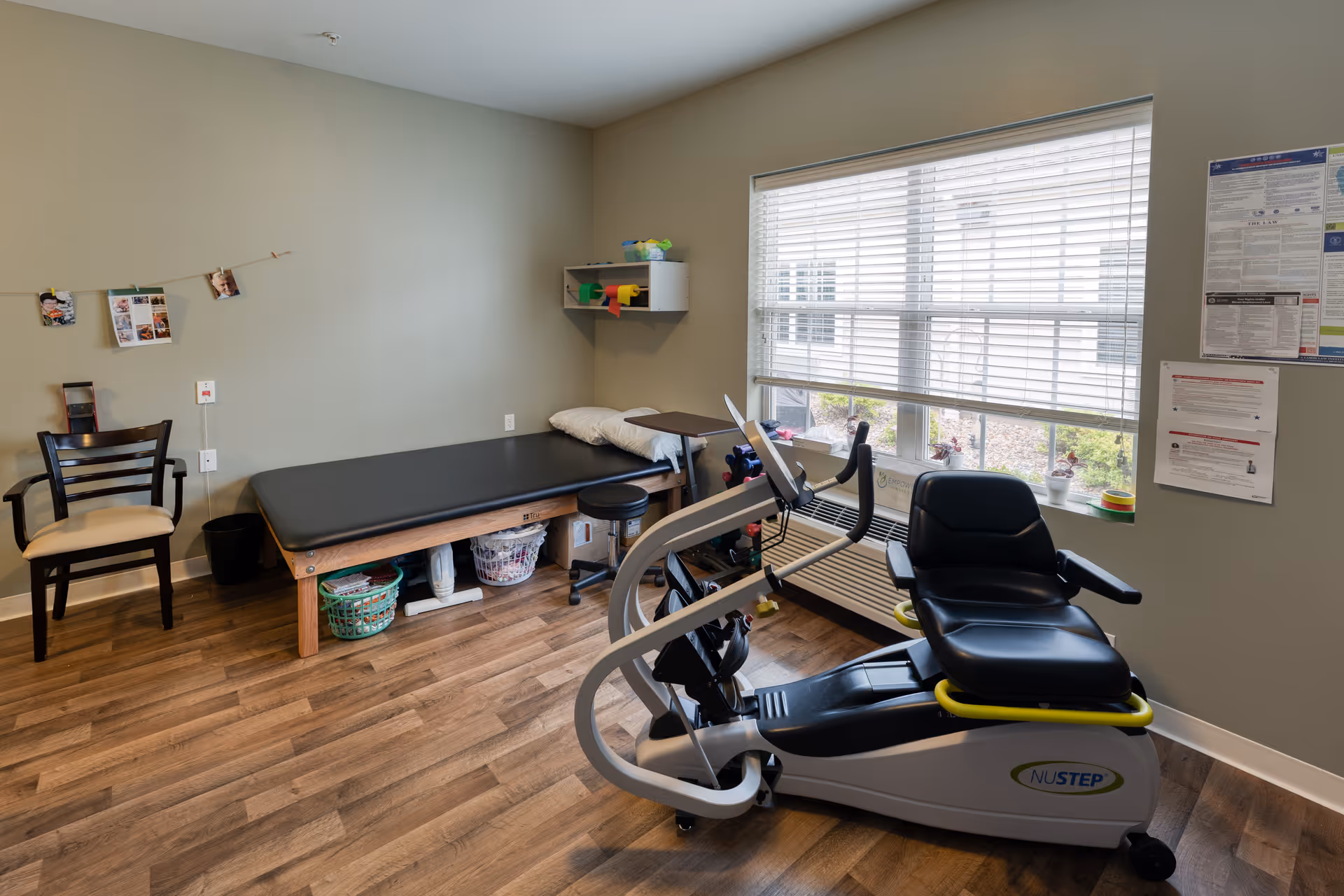Small therapy room with a recumbent exercise machine, treatment table, chair, and a large window.