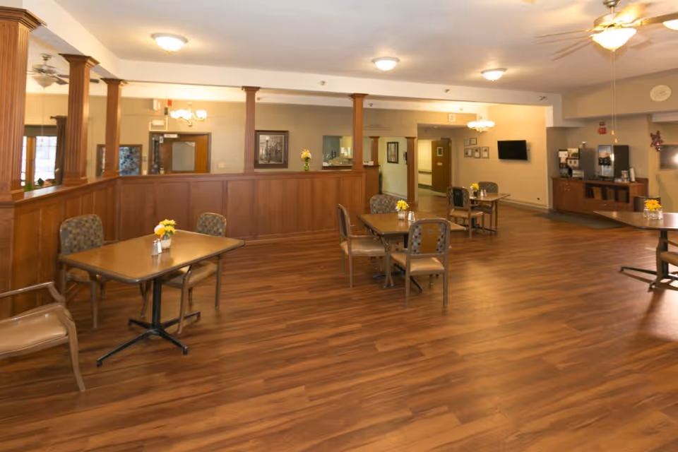 A spacious dining area with wooden floors and several tables, each with chairs and small flower arrangements. The room features wooden paneling and columns, ceiling fans, and overhead lighting. There is a beverage station with coffee machines against one wall and a flat-screen TV mounted nearby.