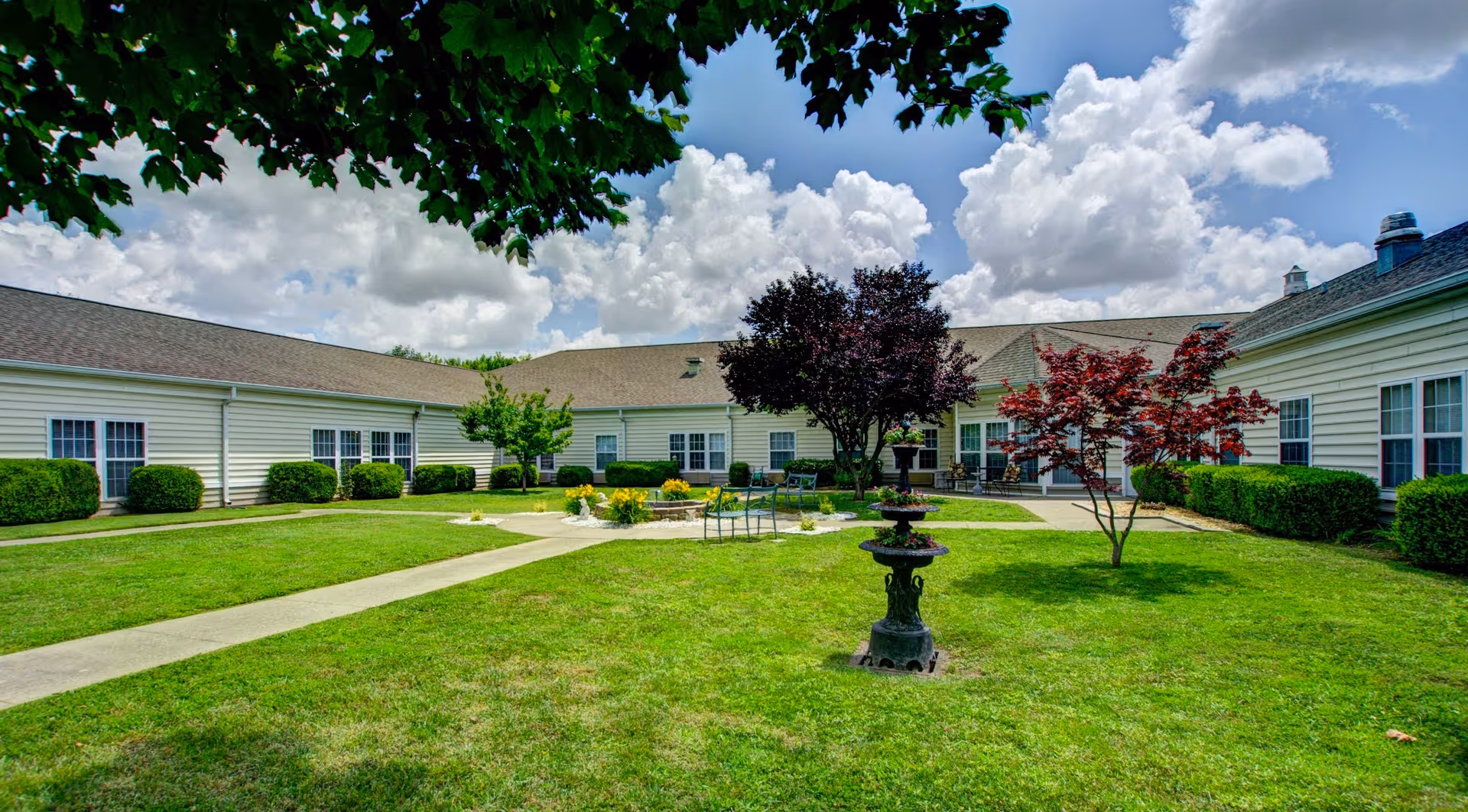 A sunny outdoor courtyard at The Bungalows at Mayfield featuring a well-maintained lawn, paved walkways, a central water fountain, several trees including a red-leafed tree, and a few chairs and tables arranged near the building. The building surrounding the courtyard has white siding and multiple windows. The sky is partly cloudy with large white clouds.