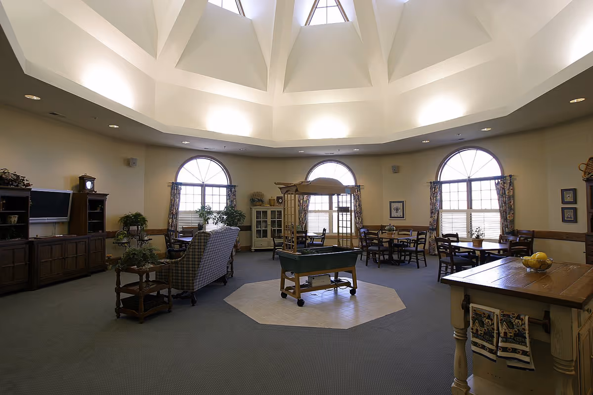 A spacious, well-lit common area with a high, vaulted ceiling and three large arched windows with floral curtains. The room features multiple seating areas with wooden chairs and tables, a plaid armchair, a wooden entertainment center with a TV, and a central decorative planter on wheels. The decor includes plants, framed pictures, and a wooden island with towels and a bowl of lemons.