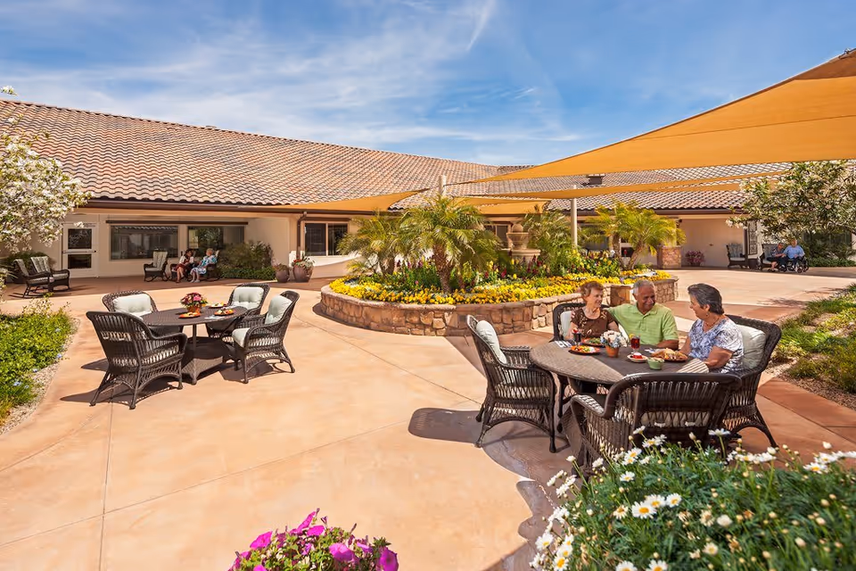 People sit and chat at wicker tables in a sunny landscaped courtyard with flower beds, a central planter, and shade sails.