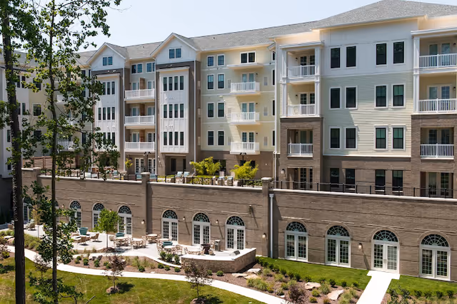Exterior view of a multi-story senior living facility with balconies, large windows, and a landscaped outdoor patio area with tables and chairs surrounded by greenery and trees.