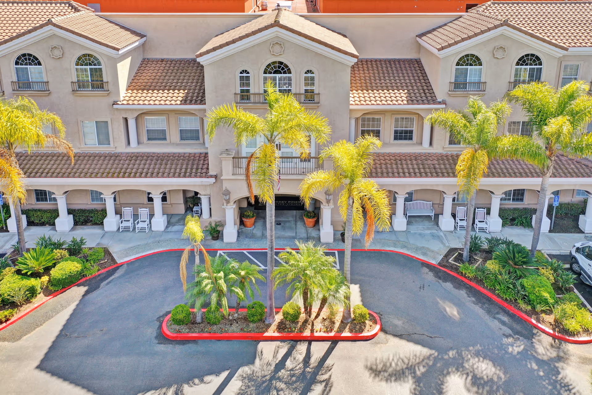 Front exterior view of a senior living facility building with beige walls and a tiled roof. The entrance is framed by columns and has potted plants on either side. Several palm trees and landscaped bushes are in front of the building, along with a driveway and parking spaces.