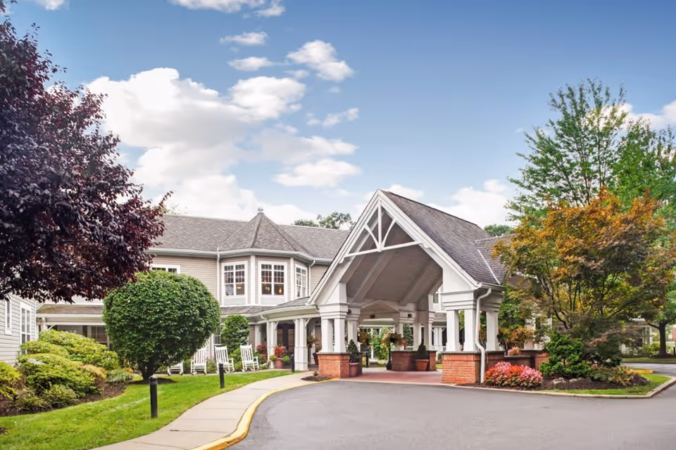 Exterior view of a senior living facility with a covered entrance, manicured bushes, trees, and a clear blue sky with some clouds.