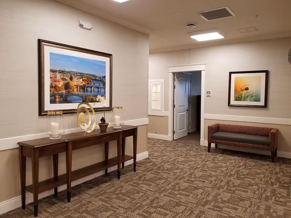 Interior hallway of a senior living facility with beige walls and patterned carpet. A wooden console table with decorative items including candles and a small plant is placed against one wall beneath a large framed picture of a cityscape with a river and bridges. On the opposite wall, there is a framed picture of a flower above a cushioned bench. An open door leads to another room.