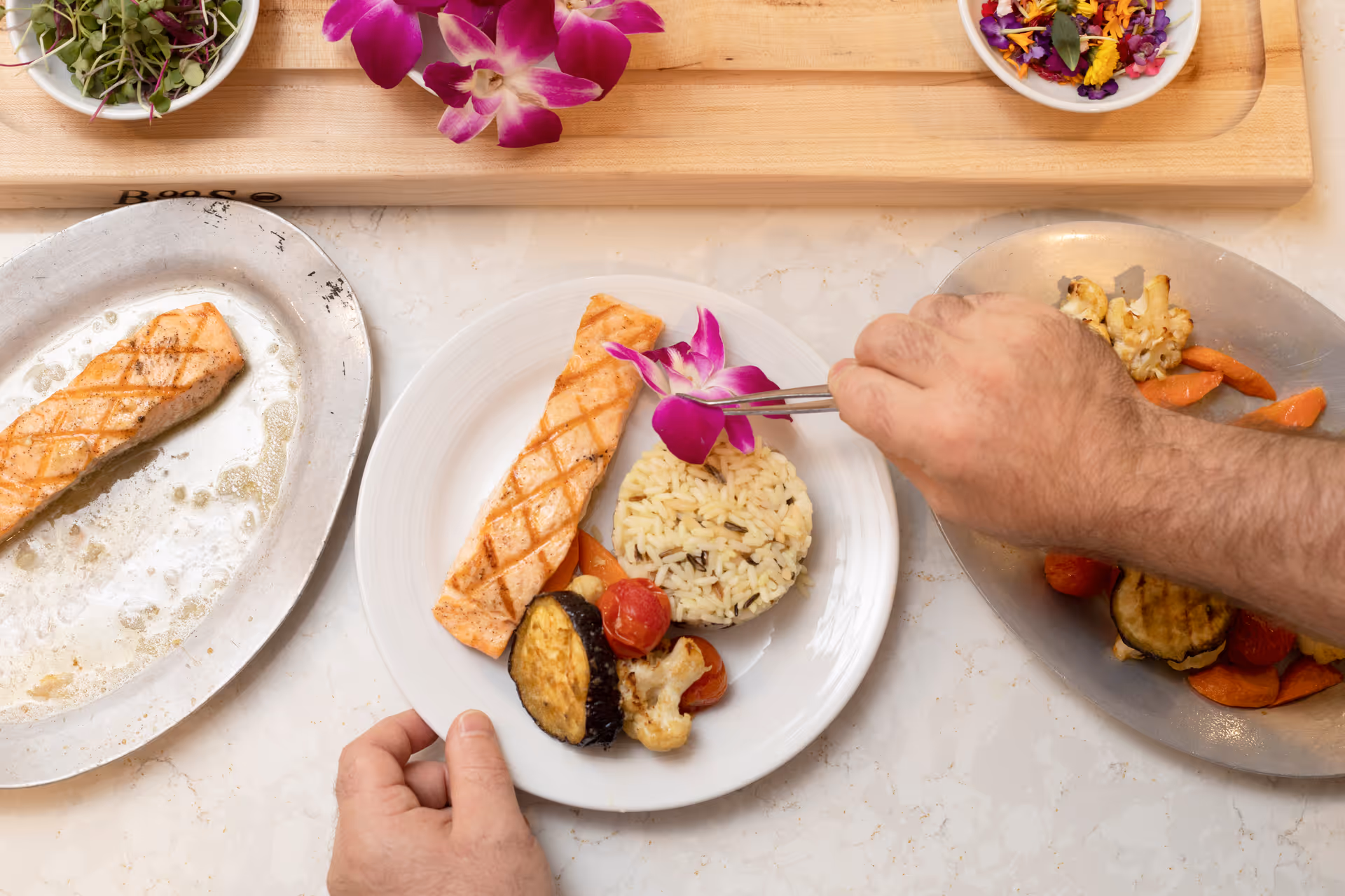 Close-up of a plated meal being garnished with a purple edible flower using tweezers. The plate contains grilled salmon, a serving of rice, and roasted vegetables including eggplant, cauliflower, and carrots. Additional plates with similar food items and bowls with greens and edible flowers are visible on a marble countertop.