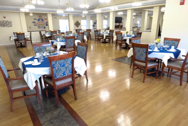 A spacious dining room with multiple tables covered in white and blue tablecloths, each set with napkins, glasses, and small floral centerpieces. The room has wooden floors, decorative chairs with blue patterned upholstery, and bright overhead lighting. Walls are adorned with some decorations and windows allowing natural light.