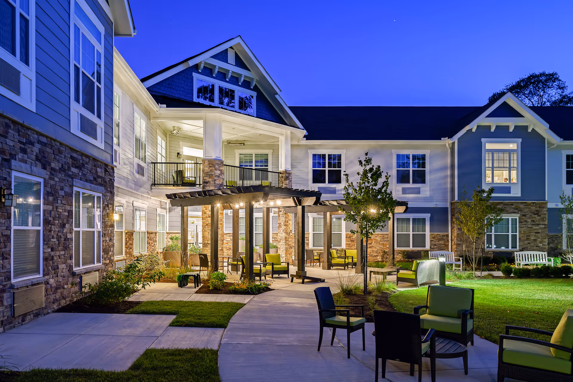Outdoor courtyard area of a senior living facility at dusk with a covered seating area, multiple chairs and tables, well-maintained landscaping, and a two-story building with stone and siding exterior in the background.