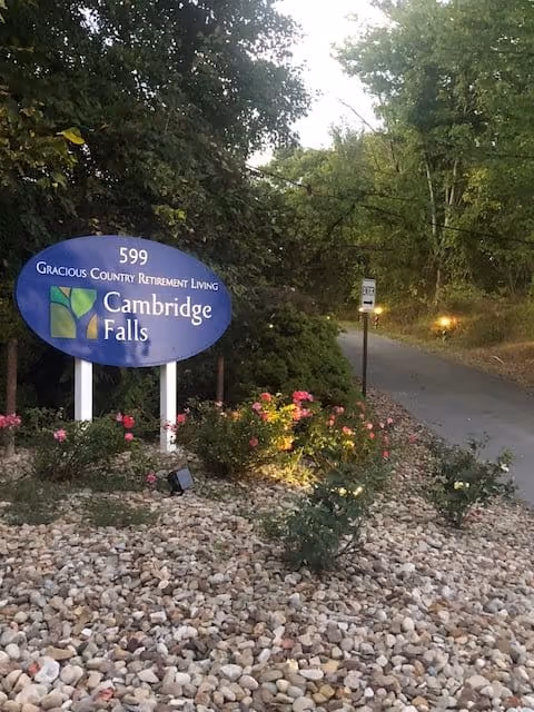 An outdoor scene showing a blue oval sign for Cambridge Falls Gracious Country Retirement Living with the number 599 above it. The sign is surrounded by small rocks and flowering plants, with a driveway and trees in the background.