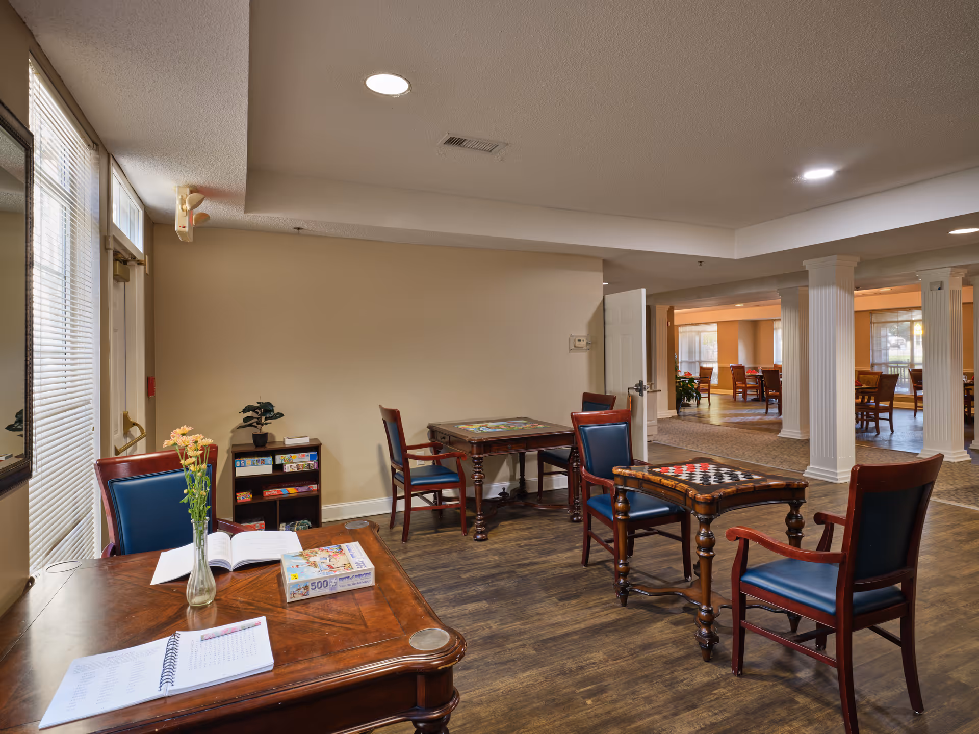 Interior view of a common area in Greenwood Place Assisted Living and Memory Care featuring several wooden tables and chairs with blue cushions. One table has a vase with flowers, an open book, and a puzzle box. Another table has a checkerboard on it. There is a small shelf with board games against the wall, and large windows with blinds letting in natural light. The room has wooden flooring and white columns separating the space.
