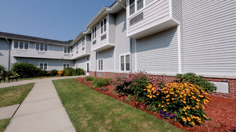 Exterior view of a light gray two-story assisted living facility building with white trim, a brick foundation, and a landscaped garden with colorful flowers along the side. A concrete walkway curves through a grassy area leading to the building entrance under a clear blue sky.