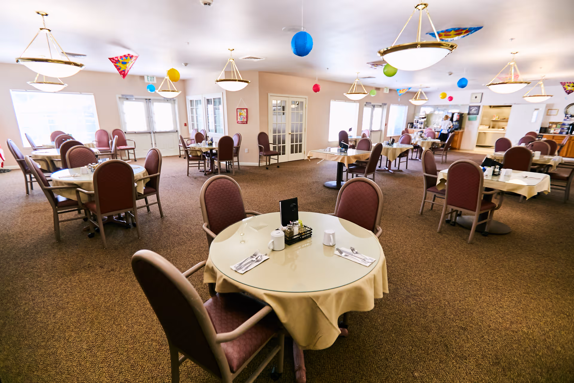 A spacious dining room with multiple round tables covered with beige tablecloths, each surrounded by four padded chairs. The room is decorated with colorful hanging paper lanterns and kites from the ceiling. There are large windows letting in natural light and a serving area in the background.