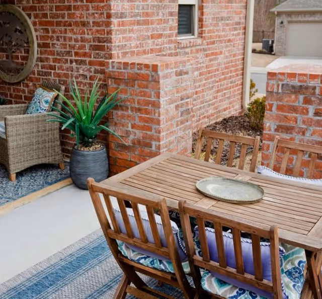 Brick-walled outdoor patio with a wooden dining table and chairs, a potted plant, and wicker seating.