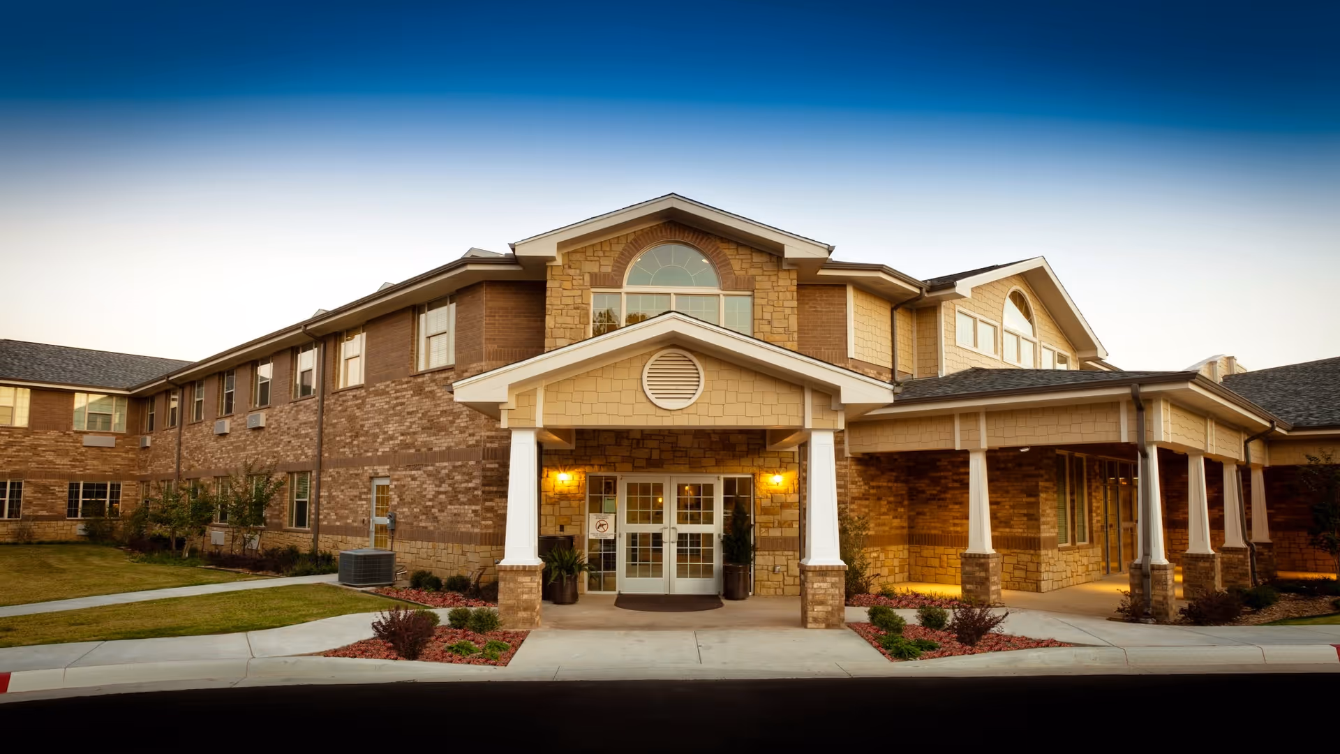 Exterior front view of a two-story senior living facility building with brick and stone facade, a covered entrance supported by white columns, landscaped greenery, and a clear sky at dusk.