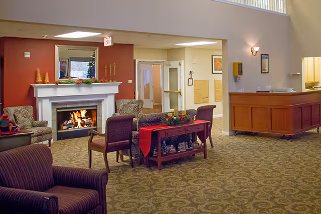 A cozy senior living facility common area with a lit fireplace, armchairs, and a small table decorated with holiday ornaments and a red runner. The room features patterned carpet, warm lighting, and a reception desk on the right side.