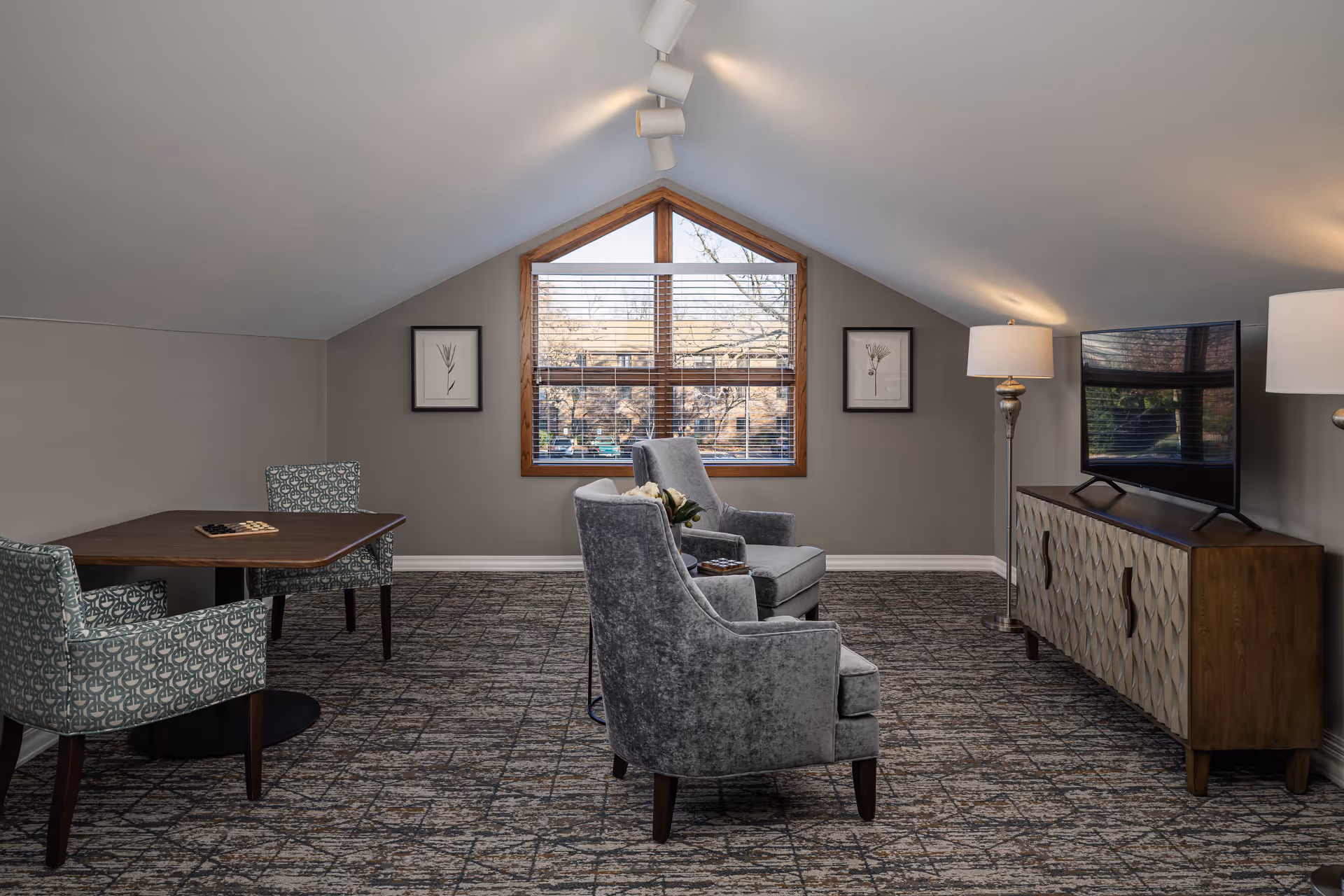 A cozy senior living community room with two gray upholstered armchairs facing a flat-screen TV on a wooden cabinet. There is a wooden table with two patterned chairs on the left side. The room has a large triangular window with blinds, two framed botanical prints on the wall, and two floor lamps providing warm lighting. The carpet has a geometric pattern, and the ceiling is sloped.