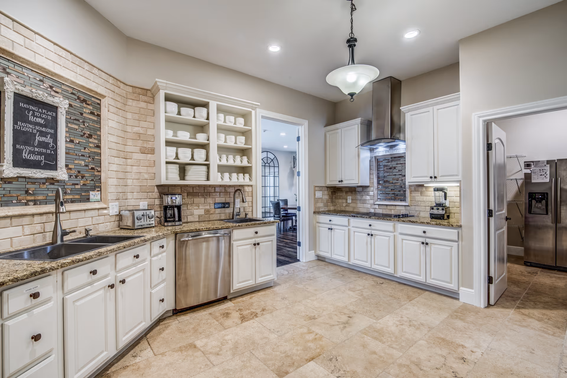 A spacious kitchen with white cabinetry, granite countertops, and beige tiled flooring. The kitchen features a double sink, dishwasher, coffee maker, toaster, and a stainless steel range hood above a stovetop. Open shelves display neatly stacked white dishes and bowls. There is a decorative framed sign on the brick backsplash that reads, 'Having a place to go is home. Having someone to love is family. Having both is a blessing.' Two doorways lead to other rooms, one showing a dining area and the other a room with a stainless steel refrigerator and shelving.