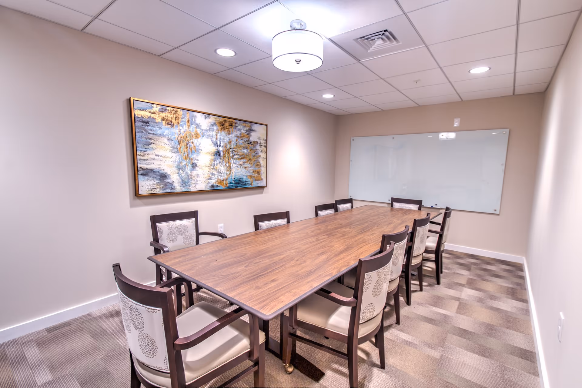 A small conference room with a long wooden table surrounded by chairs, wall art, and a whiteboard.