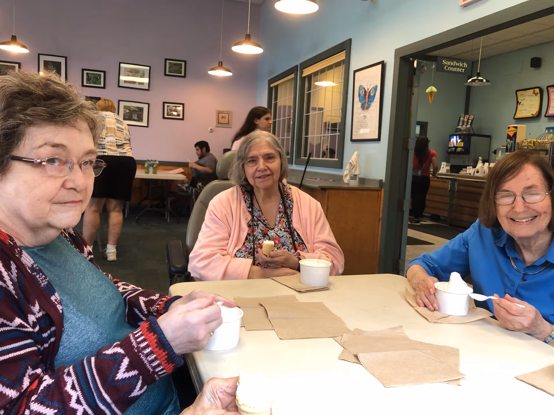 Three elderly women sitting around a table enjoying ice cream in a communal dining or activity area. The room has light blue walls with framed pictures, pendant lights hanging from the ceiling, and a sign indicating a Sandwich Counter in the background where other people are present.