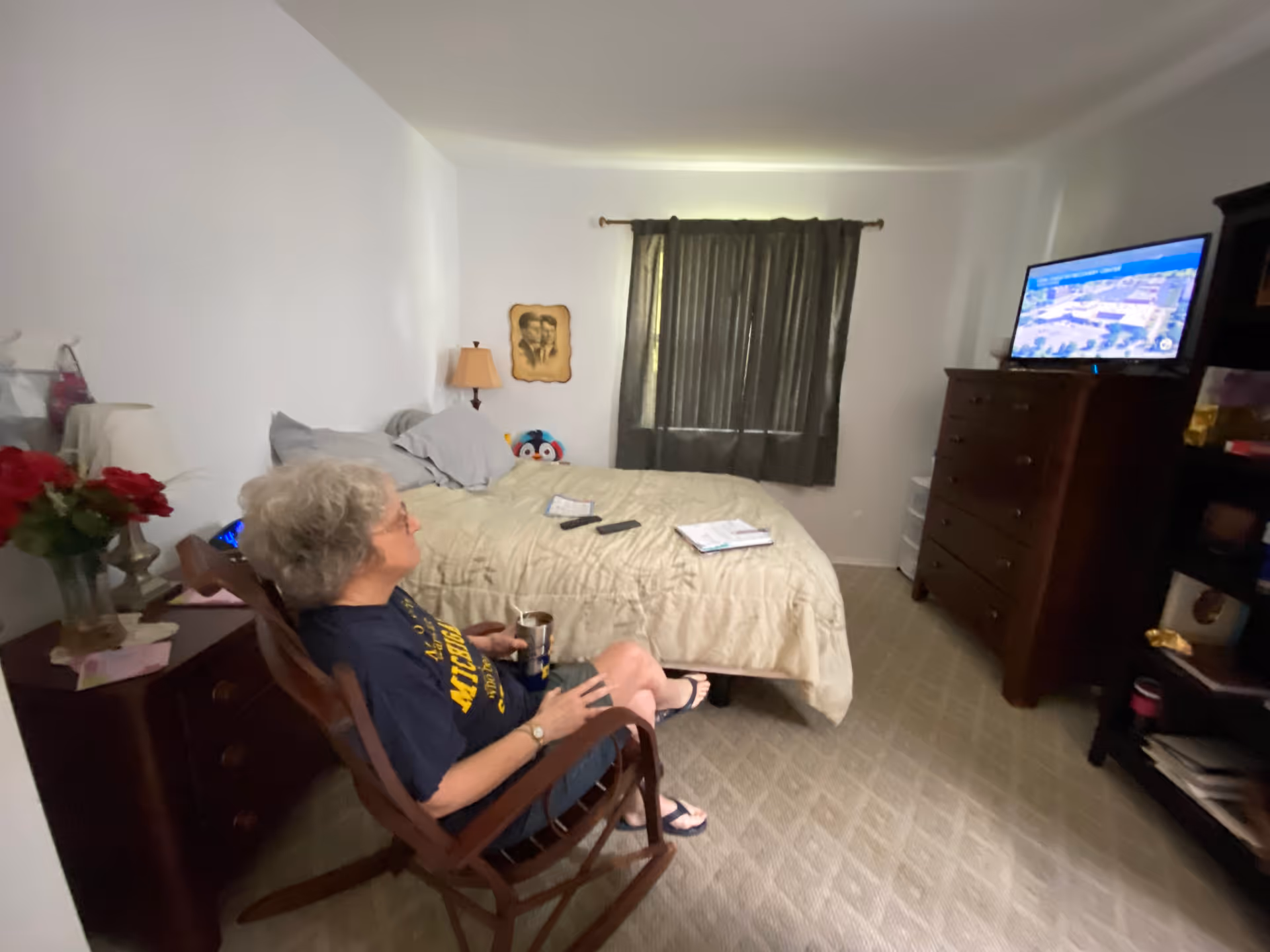 An elderly woman with gray hair sits in a wooden rocking chair in a bedroom. She is holding a cup and watching a television mounted on a dresser. The room has a bed with beige bedding, a nightstand with a lamp and flowers, a window with dark curtains, and a bookshelf with various items.