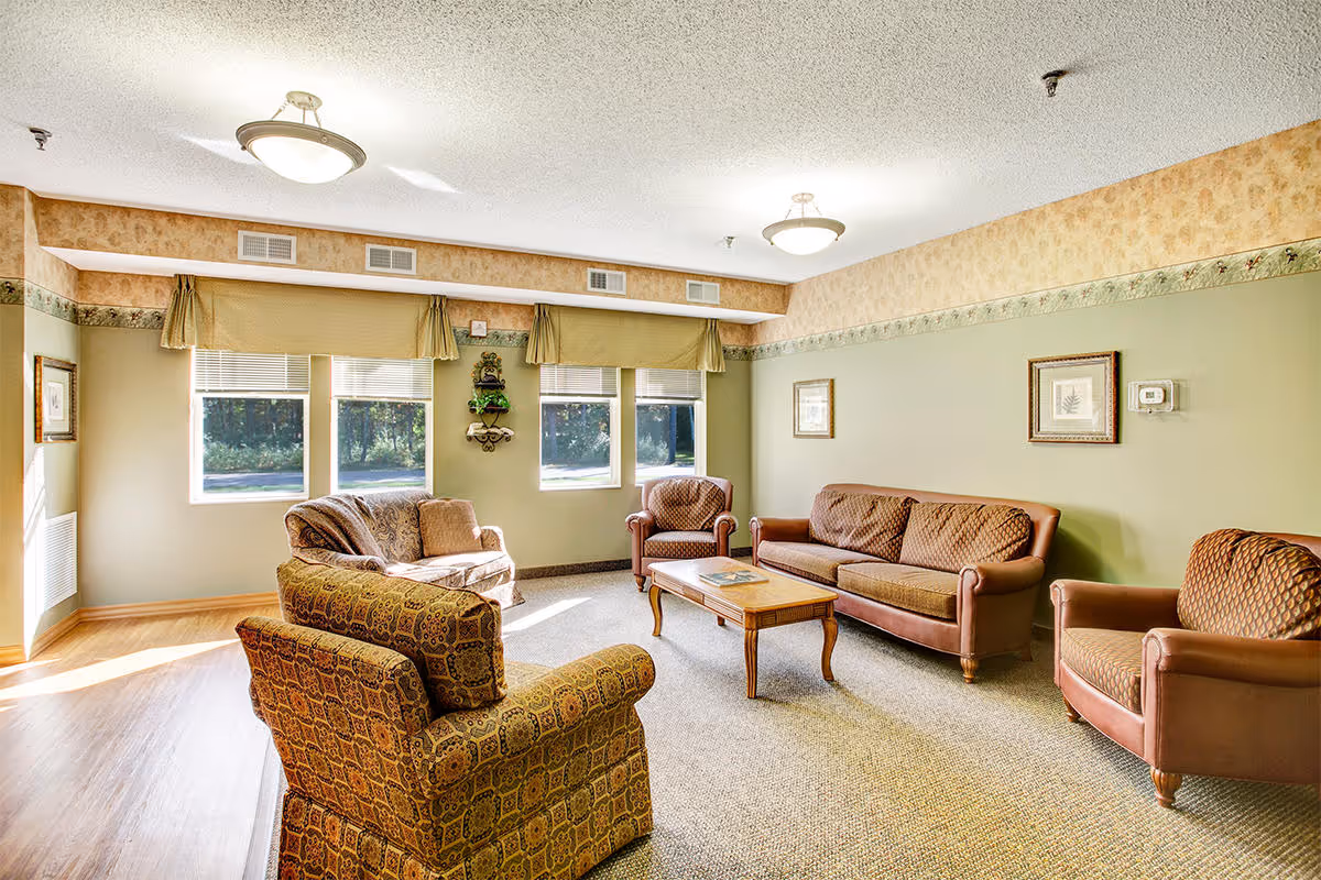 A cozy living room with a mix of patterned and solid brown upholstered chairs and sofas arranged around a wooden coffee table. The room has green walls with a decorative border near the ceiling, three windows with beige valances letting in natural light, and framed artwork on the walls. The floor is carpeted with a section of wood flooring near the windows.