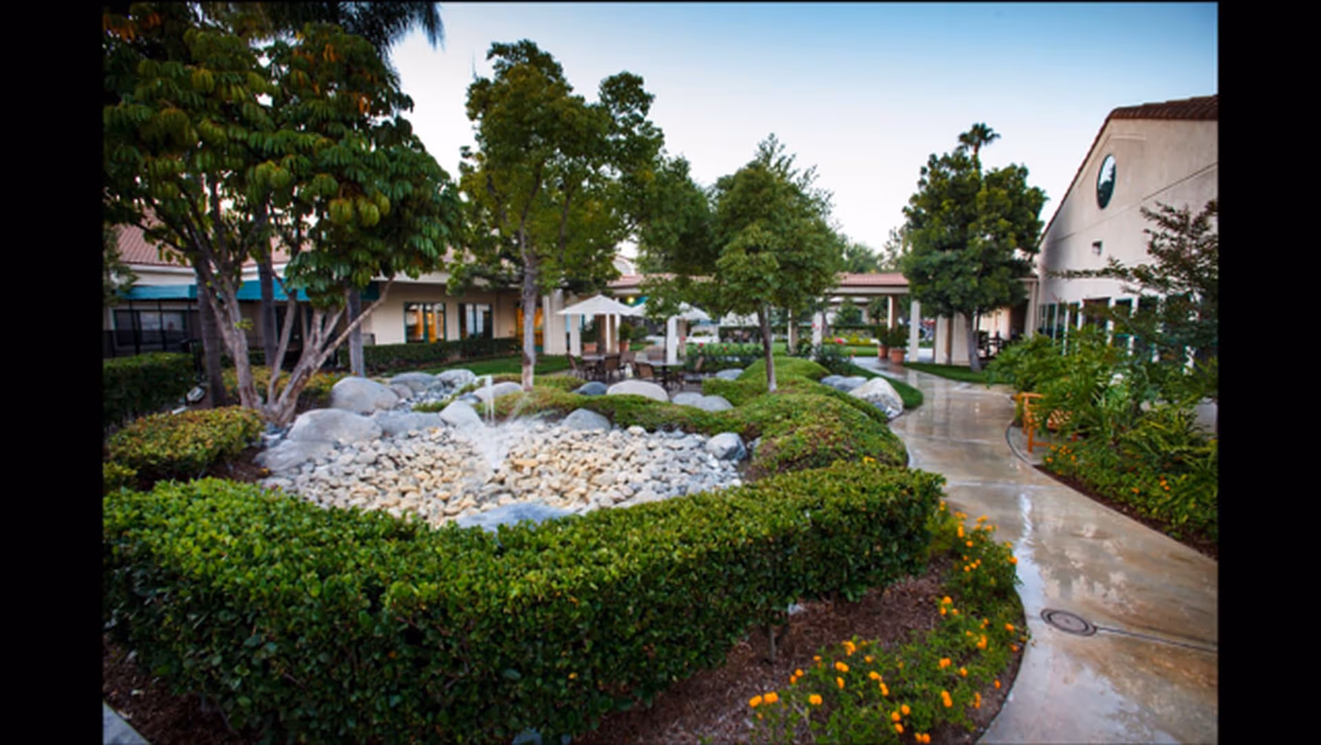 Outdoor garden area at Park Vista Assisted Living and Memory Care featuring a small rock fountain surrounded by neatly trimmed hedges, trees, and flowering plants, with a paved walkway curving around the garden and buildings visible in the background.