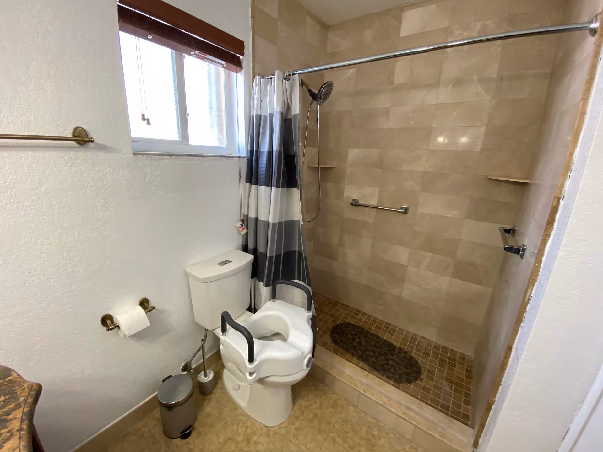 Accessible bathroom with a raised toilet seat and armrests next to a tiled walk-in shower with a striped curtain.