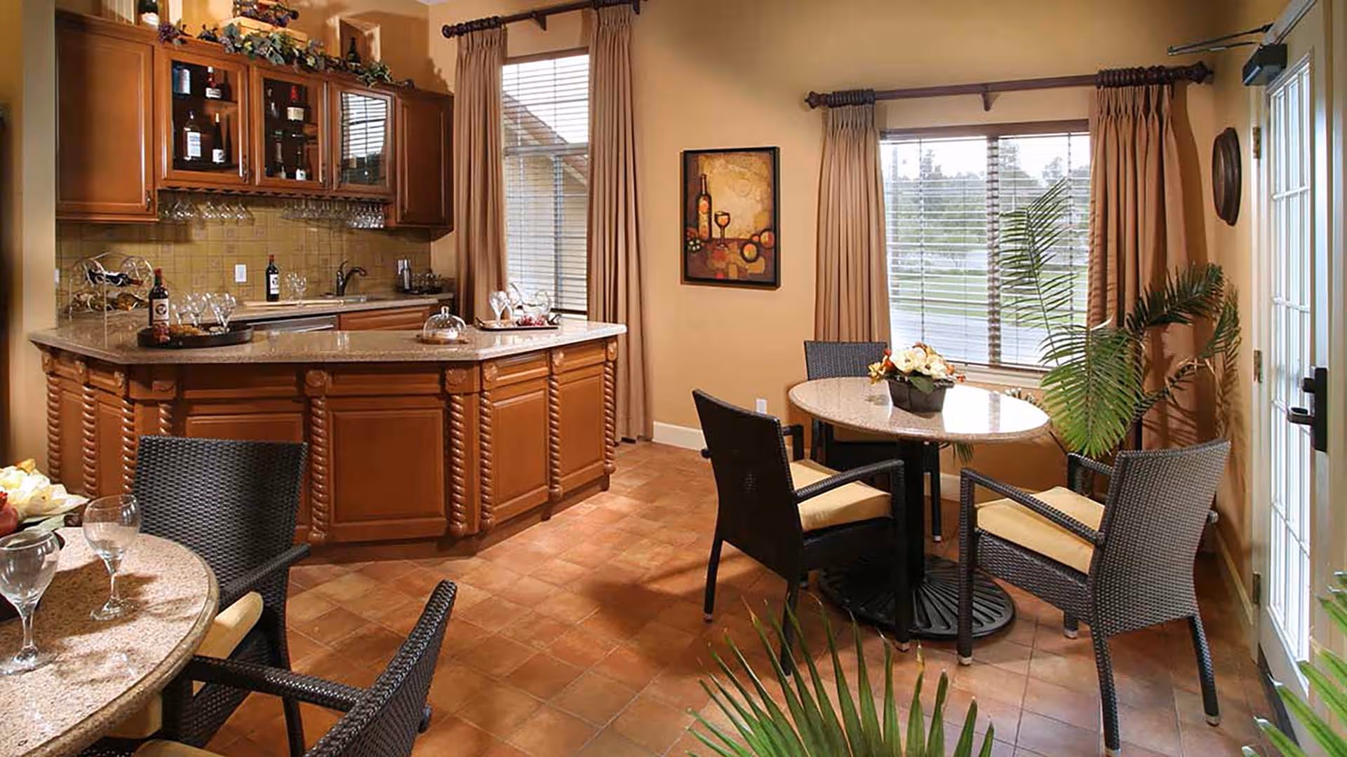 A cozy kitchen and dining area with wooden cabinetry, a granite countertop island, and two round dining tables with wicker chairs. The room has large windows with beige curtains, a framed painting on the wall, and some decorative plants.
