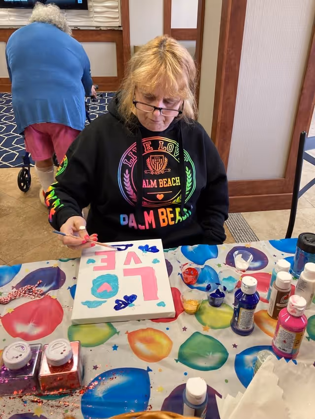 A woman wearing glasses and a black hoodie with colorful text 'Palm Beach' is painting a canvas with the word 'LOVE' in pink letters and blue hearts. She is seated at a table covered with a colorful balloon-patterned tablecloth, surrounded by various paint bottles and art supplies. In the background, an elderly person with white hair, wearing a blue top and pink shorts, is walking with a walker in a room with tiled floors and wooden paneling.