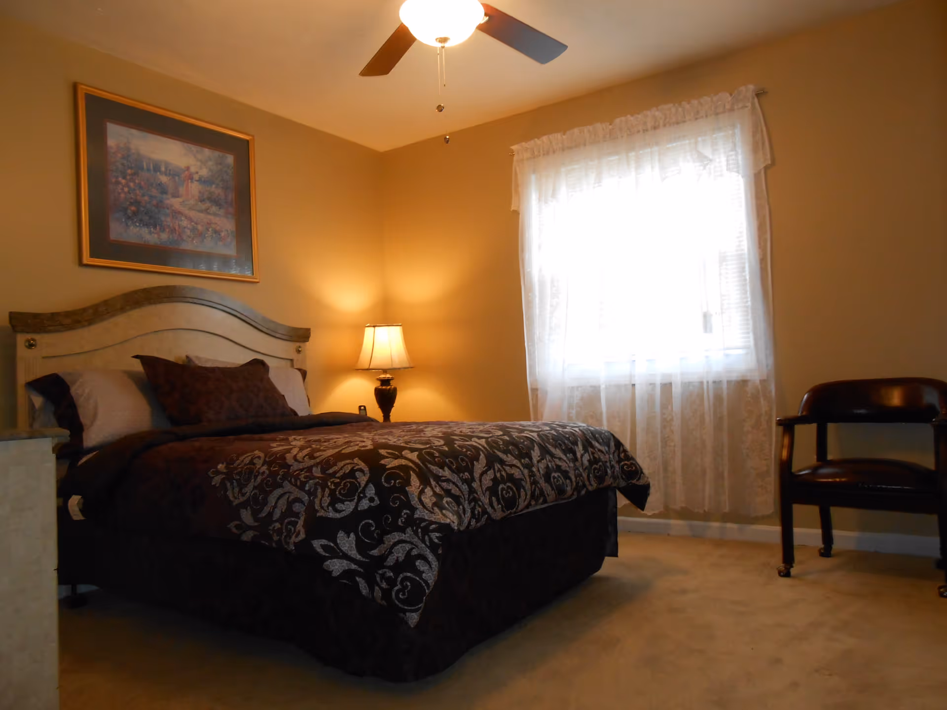 A bedroom with a large bed covered in a dark patterned comforter and pillows, a wooden headboard, a bedside table with a lamp, a window with sheer white curtains, a ceiling fan, a framed painting on the wall, and a wooden chair with armrests on the right side.